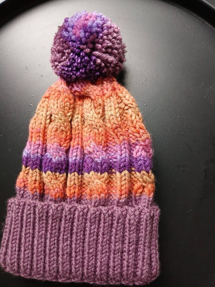 Cable-knit bobble hat in orange and peach with violet and pink bands, a mauve cuff and a purple bobble